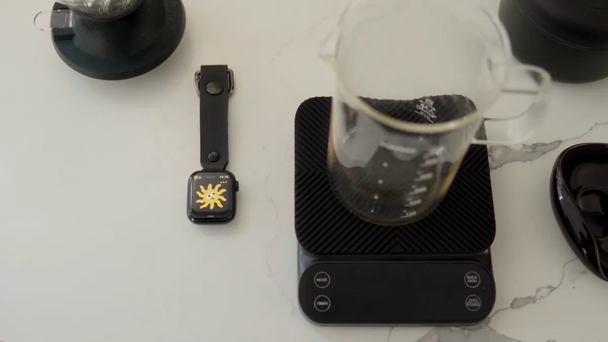 The watch on its strap loop on a marble counter, next to a coffee scale and a carafe. Ray is thinking on the screen.