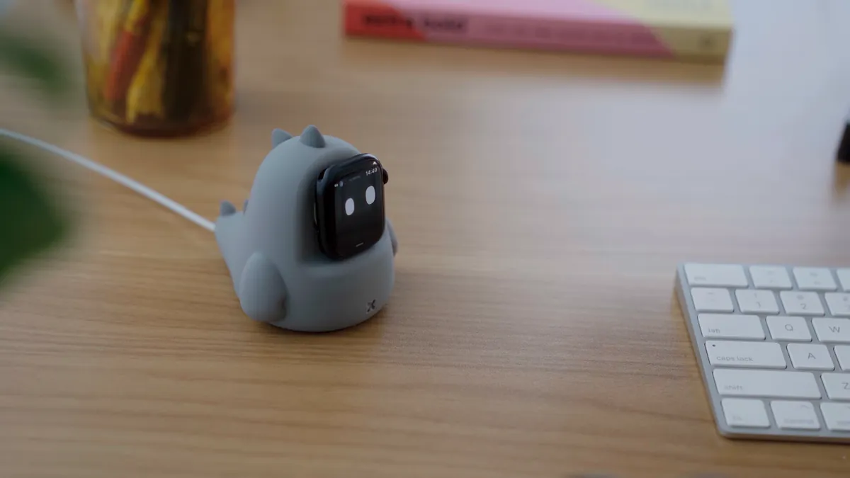 The watch on a small grey dinosaur charging stand on a wooden desk, next to a keyboard. Peeps is on the screen.